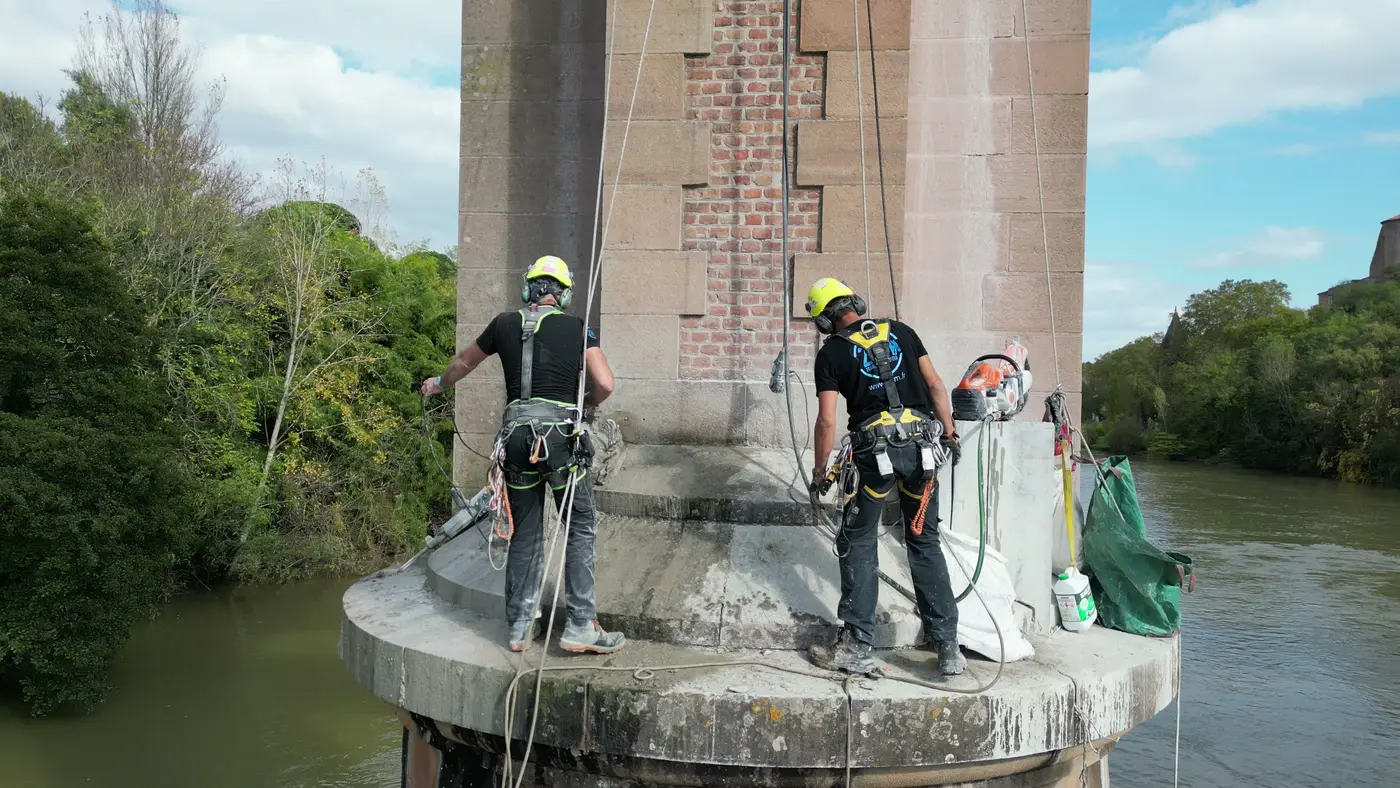Deux cordistes en suspension sur l'avant-bec d'une pile, sacs à gravats et outillage, au-dessus du Tarn